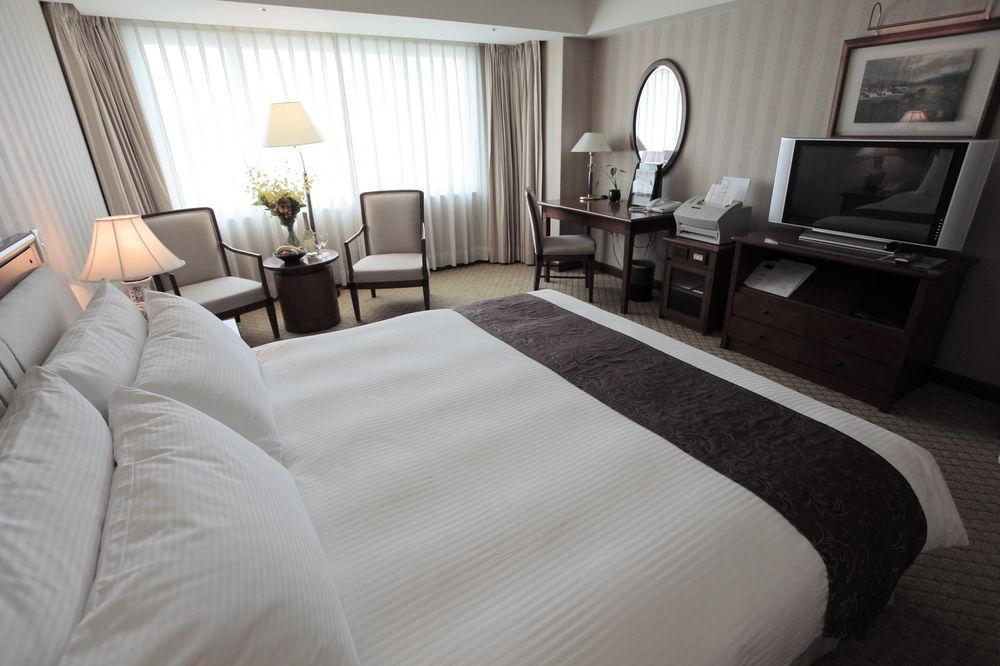 Evergreen Laurel Hotel - Taichung Bagian luar foto The photo shows a well-furnished hotel room. In the foreground, there is a large bed with white bedding and a dark-colored throw at the foot. Beside the bed, there is a bedside lamp. 

In the background, natural light filters through large windows co