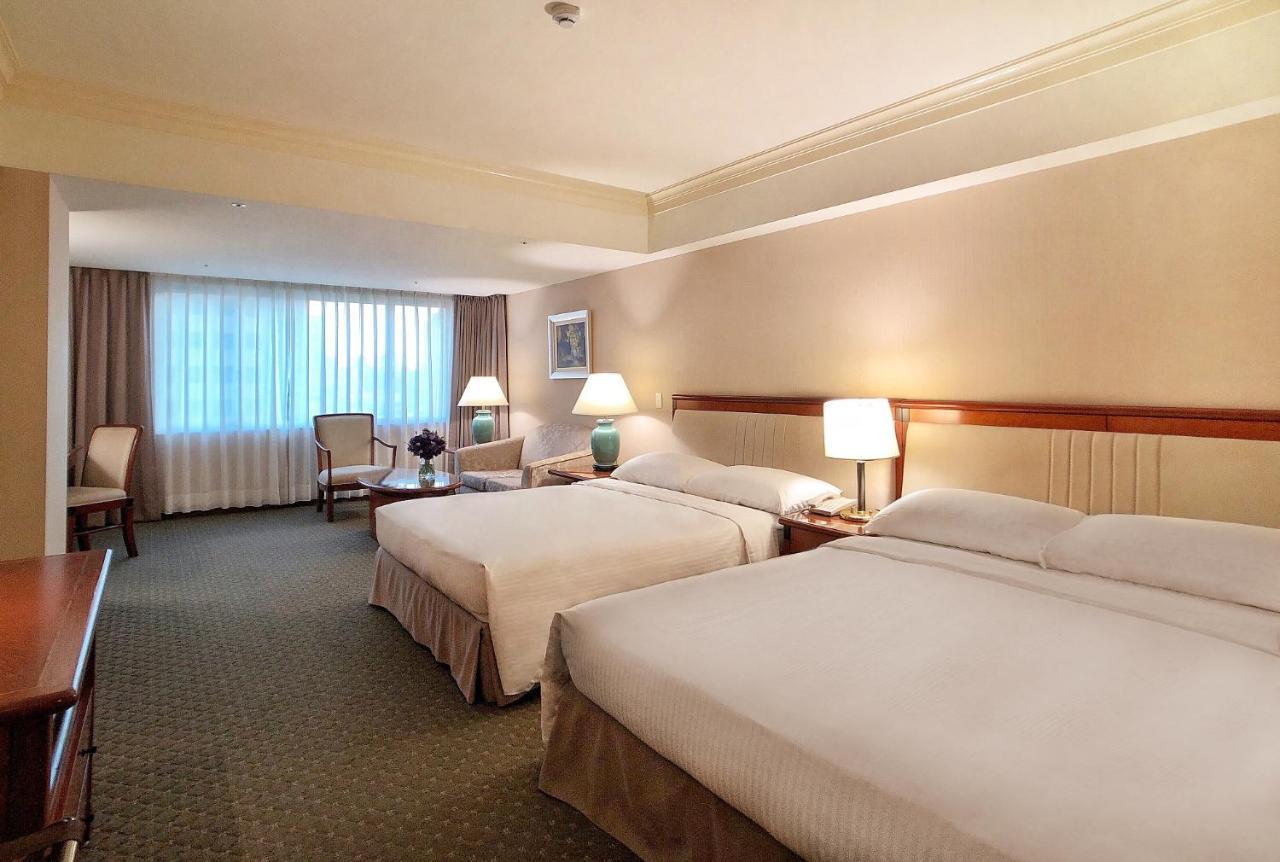 Evergreen Laurel Hotel - Taichung Bagian luar foto The photo shows a hotel room featuring two queen-sized beds with white linens. The room is well-lit and has a warm, neutral color palette. There are bedside lamps on each side of the beds and a small table. To the side, there's a comfortable armchair