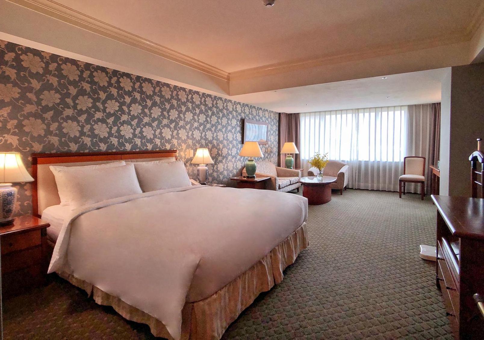 Evergreen Laurel Hotel - Taichung Bagian luar foto The photo shows a hotel room with a spacious and comfortable layout. It features a large bed with white bedding, placed against a decorative wallpapered wall. On either side of the bed, there are lamps providing soft lighting. In addition to the bed,