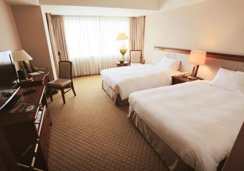 Evergreen Laurel Hotel - Taichung Bagian luar foto The photo shows a hotel room featuring two twin beds with white bedding. There's a chair positioned near one of the beds and a desk with a computer setup against one wall. Large windows allow natural light to enter the room, and light-colored curtain