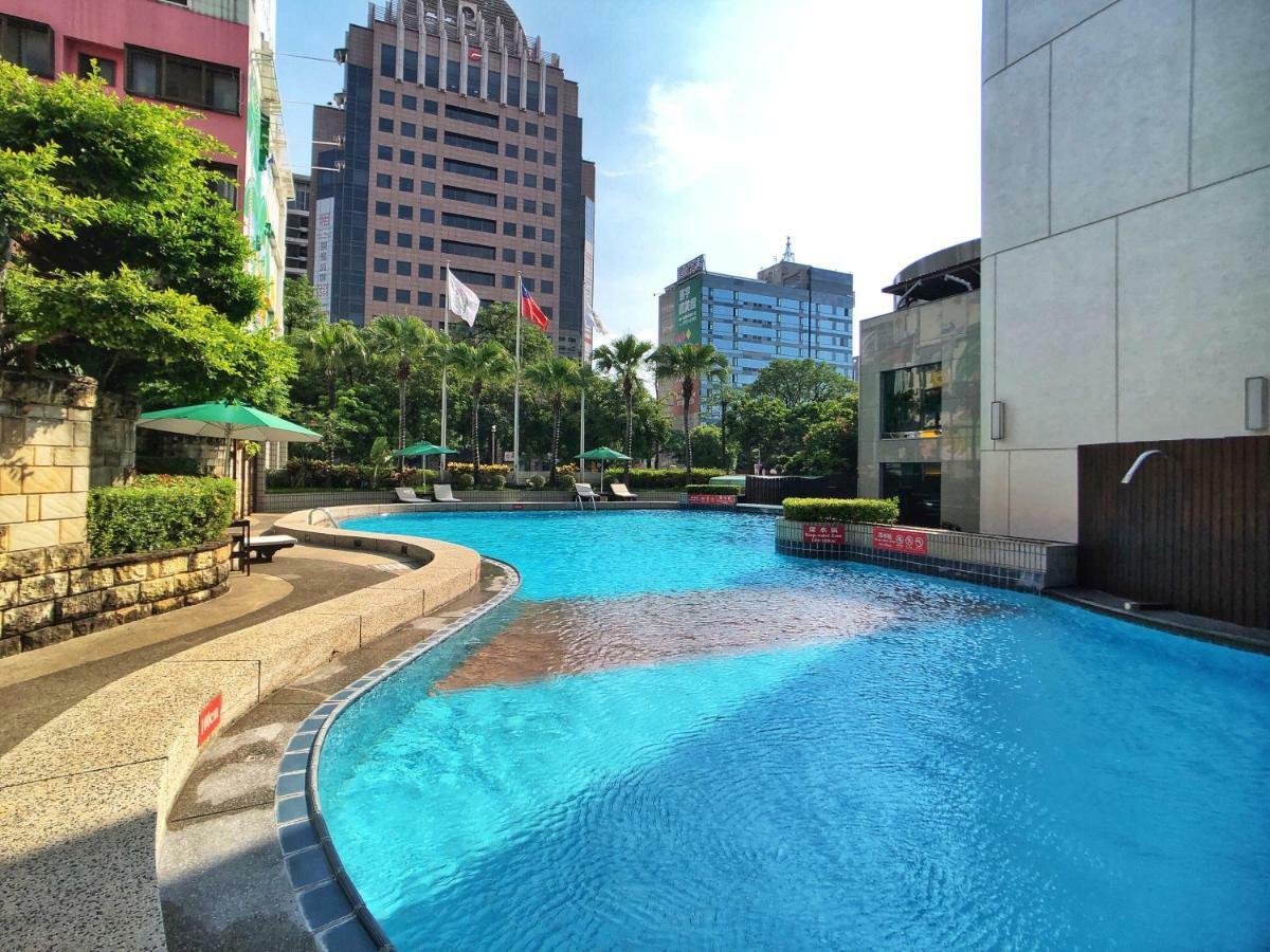 Evergreen Laurel Hotel - Taichung Bagian luar foto The photo shows a swimming pool set in a modern urban environment. The pool has a curved edge and is surrounded by greenery, including trees and umbrellas for shade. In the background, there are several tall buildings, indicating a cityscape, with so
