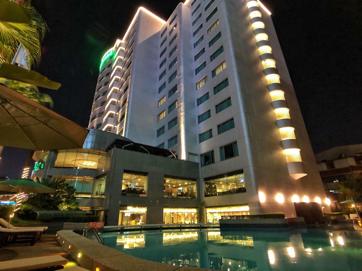 Evergreen Laurel Hotel - Taichung Bagian luar foto The photo shows a tall hotel building at night, illuminated by lights. The architecture appears modern, with several floors and large windows. In the foreground, there is a swimming pool that reflects the lights from the building, creating a serene a