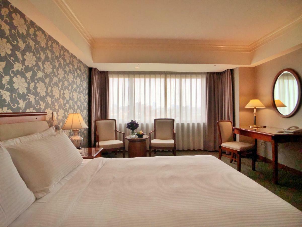 Evergreen Laurel Hotel - Taichung Bagian luar foto The photo shows a hotel room with a comfortable and inviting design. There is a large bed with crisp white bedding on the left side of the image. To the right, there are two elegant chairs with a small round table between them, adorned with a bouquet
