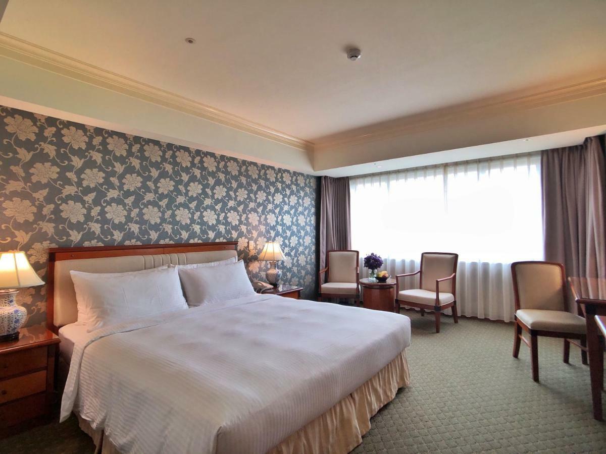 Evergreen Laurel Hotel - Taichung Bagian luar foto The photo shows a hotel room that features a large bed with a white bedspread positioned against a decorative floral wallpaper. There are two bedside lamps on wooden nightstands flanking the bed. To the side of the room, there are two chairs and a sm