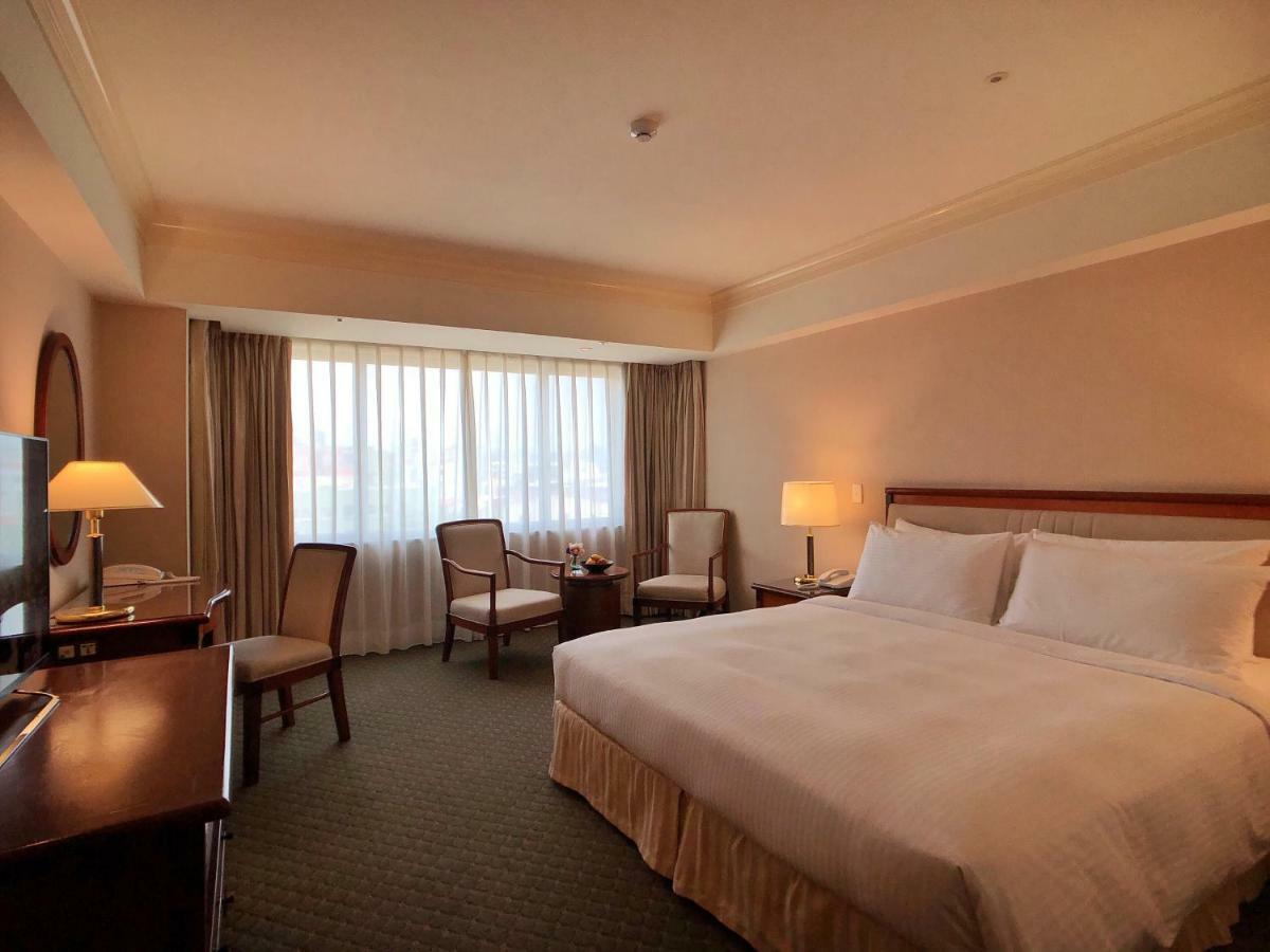Evergreen Laurel Hotel - Taichung Bagian luar foto The photo shows a hotel room featuring a large bed with white linens, a wooden bedside table with a lamp, and a flat-screen television. There are two chairs with a small table next to them positioned by a large window, which has sheer curtains lettin