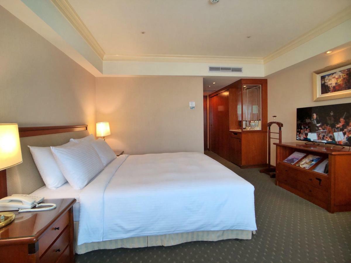 Evergreen Laurel Hotel - Taichung Bagian luar foto The photo shows a hotel room featuring a comfortable bed with white linens and pillows, positioned against a light-colored wall. There is a wooden bedside table with a lamp and an open book next to the bed. On the opposite side, there’s a large flat-