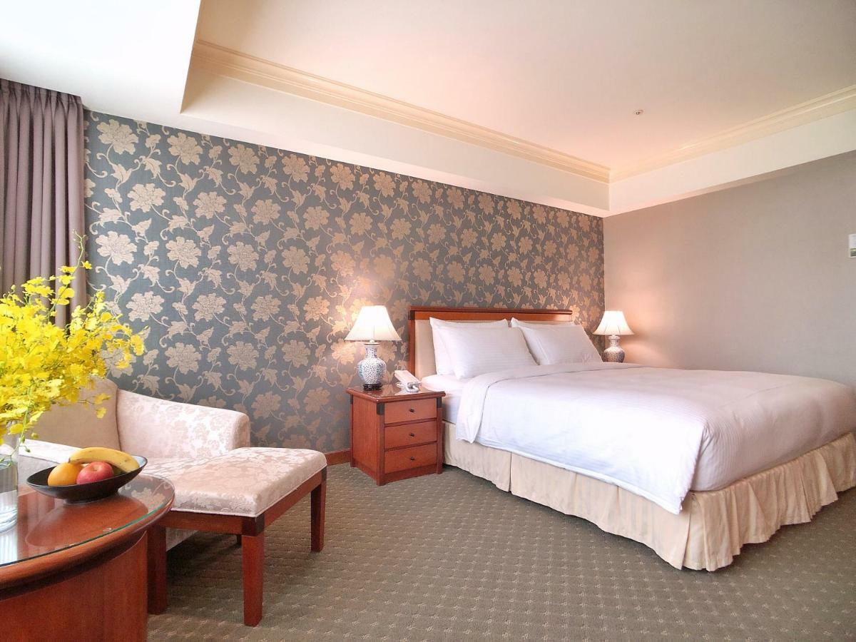 Evergreen Laurel Hotel - Taichung Bagian luar foto The photo shows a well-decorated hotel room. It features a large bed with white bedding, situated against a patterned wall with floral designs. On either side of the bed are bedside tables with lamps. There is a small seating area that includes a cha