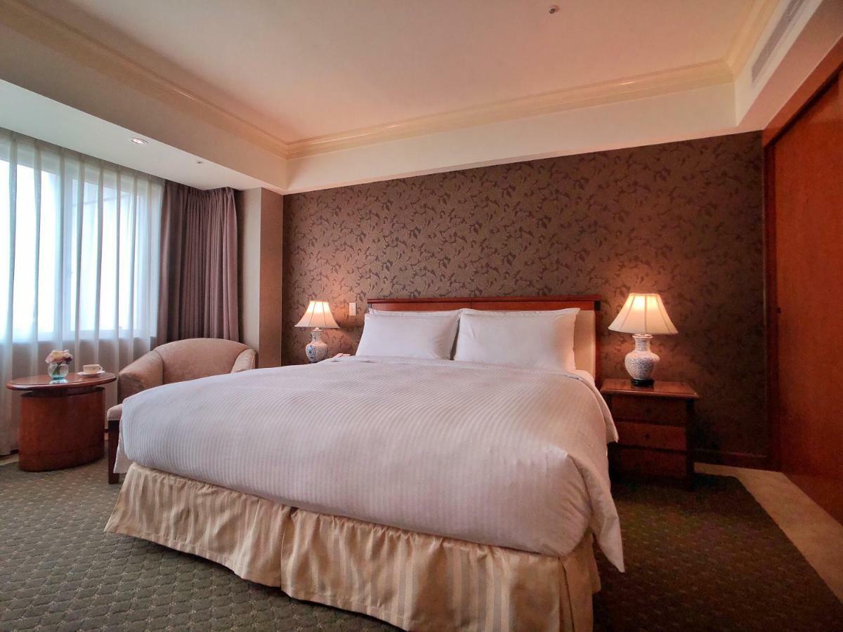 Evergreen Laurel Hotel - Taichung Bagian luar foto The photo shows a neatly arranged hotel room featuring a large bed with white linens and a light-colored bed skirt. On either side of the bed, there are small wooden nightstands with lamps. To the left of the bed, there's a comfortable chair and a ro