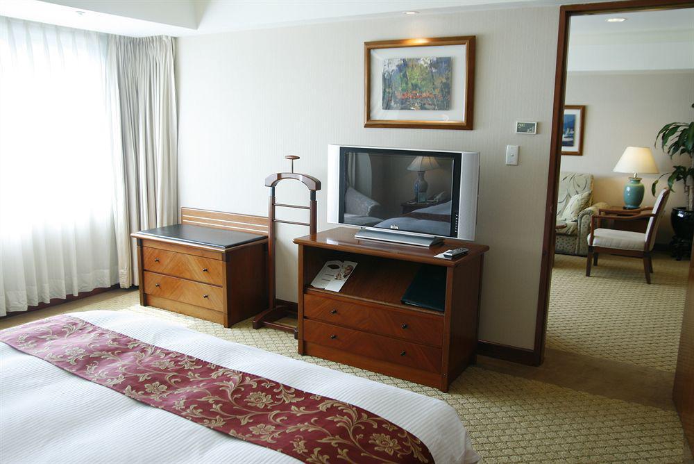 Evergreen Laurel Hotel - Taichung Bagian luar foto The photo depicts a hotel room interior. In the foreground, there is a neatly made bed with a decorative red bedspread. To the left, a large window allows natural light to fill the room, with light-colored curtains drawn to one side. 

Next to the be