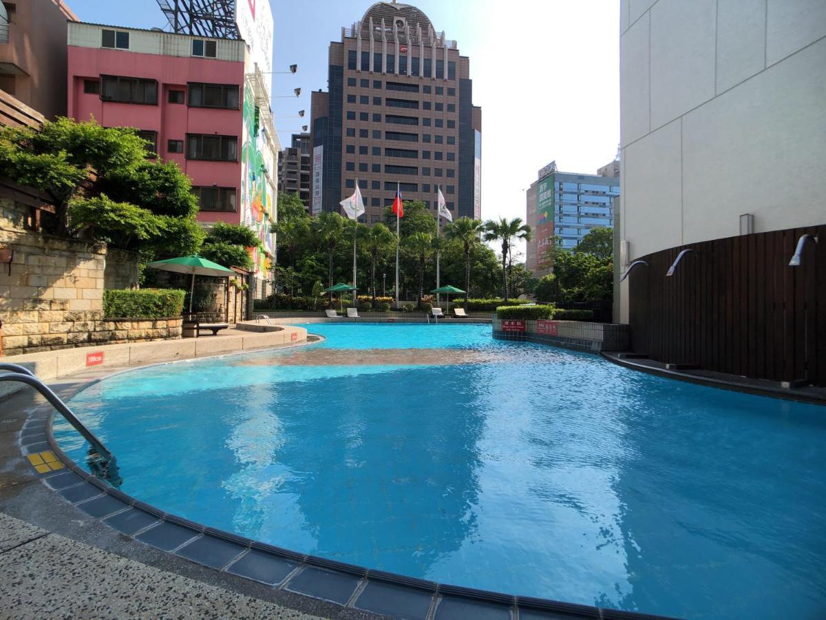 Evergreen Laurel Hotel - Taichung Bagian luar foto The photo shows a swimming pool area set in an outdoor environment. The pool has a clear blue water and a gently curving shape. Surrounding the pool are some green trees and plants, along with a few umbrellas providing shade. In the background, there