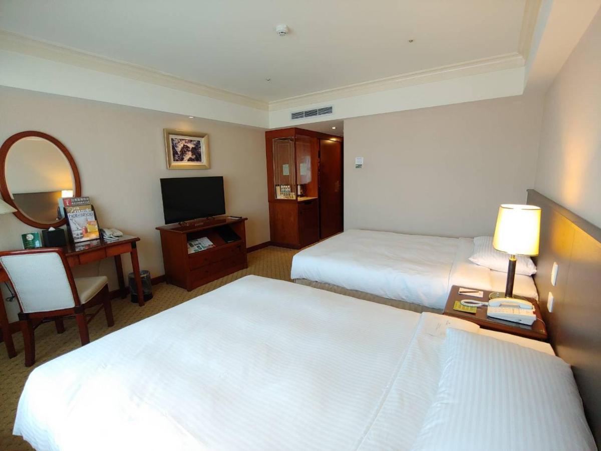 Evergreen Laurel Hotel - Taichung Bagian luar foto The photo shows a hotel room with two beds, neatly made with white linens. There is a desk and chair in one corner, accompanied by a flat-screen TV mounted on the wall. A large mirror and a wooden cabinet are also visible. The room is illuminated by 